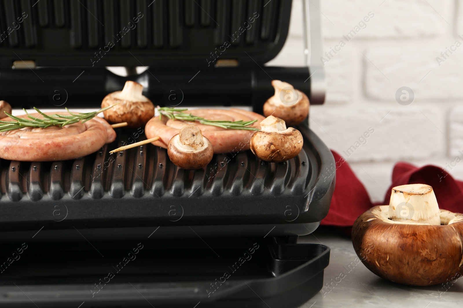 Photo of Electric grill with homemade sausages, rosemary and mushrooms on light table, closeup