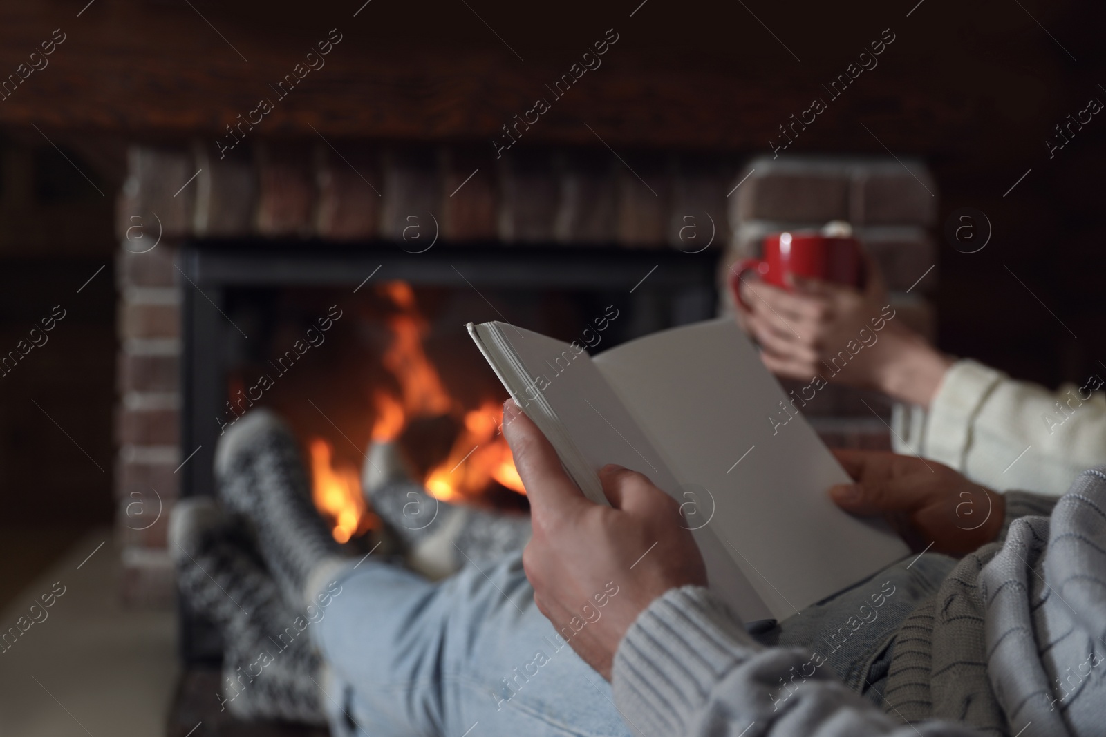 Photo of Man reading book and his girlfriend near burning fireplace at home, closeup