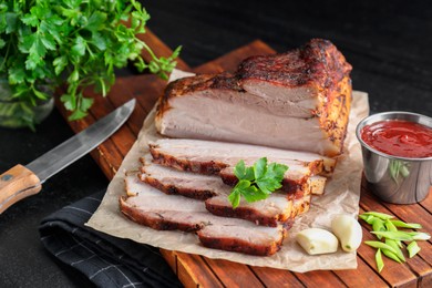 Pieces of baked pork belly served with sauce and parsley on black textured table, closeup