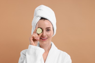 Photo of Beautiful woman in bathrobe covering eye with piece of cucumber on beige background