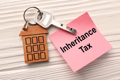 Photo of Inheritance Tax. Paper note and key with key chain in shape of house on white wooden table, flat lay