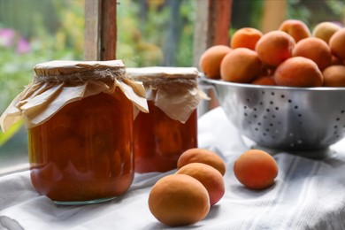 Photo of Delicious ripe apricots with jars of homemade jam on table near window