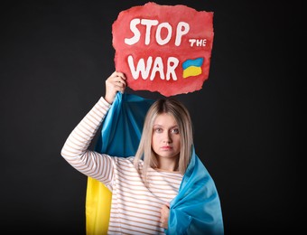 Photo of Sad woman wrapped in Ukrainian flag holding poster with words Stop the War on black background