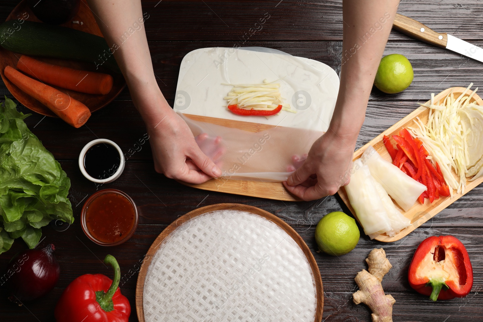 Photo of Making delicious spring rolls. Woman wrapping fresh vegetables into rice paper at wooden table, flat lay