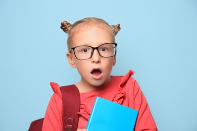 Photo of Cute little girl with glasses, backpack and textbook on light blue background