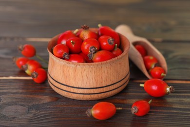 Photo of Ripe rose hip berries with bowl and scoop on wooden table, closeup