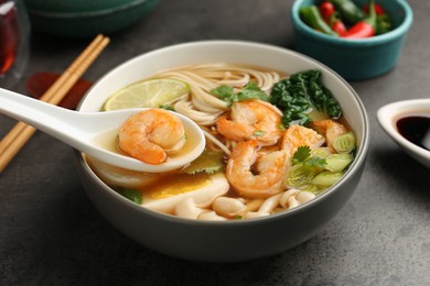 Photo of Eating delicious ramen with shrimps from bowl with spoon at grey table, closeup. Noodle soup