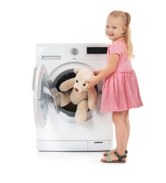 Photo of Cute little girl taking teddy bear out of washing machine on white background. Laundry day