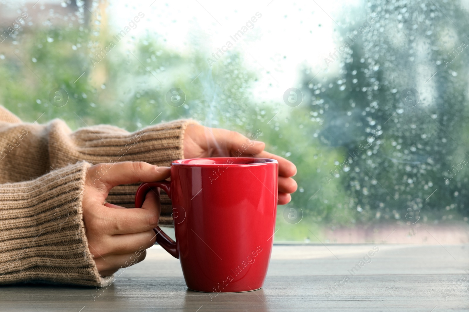 Photo of Woman with cup of hot drink at wooden table near window on rainy day, closeup