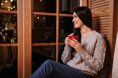 Beautiful woman with cup of coffee near window indoors. Christmas celebration