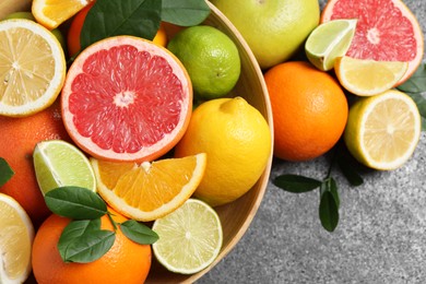 Photo of Different fresh citrus fruits and leaves in bowl on grey textured table, flat lay