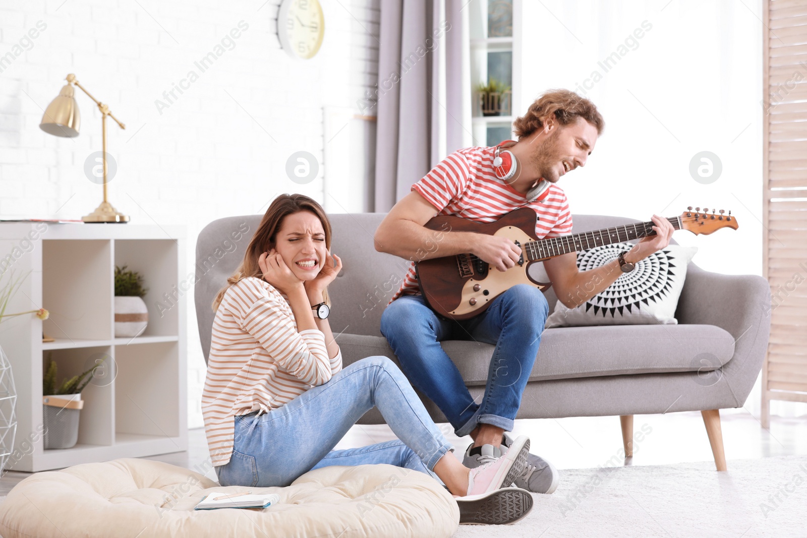 Photo of Young man playing electric guitar badly for displeased girlfriend in living room. Talentless musician