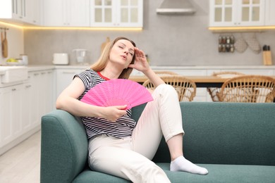 Photo of Woman waving pink hand fan to cool herself on sofa at home. Space for text