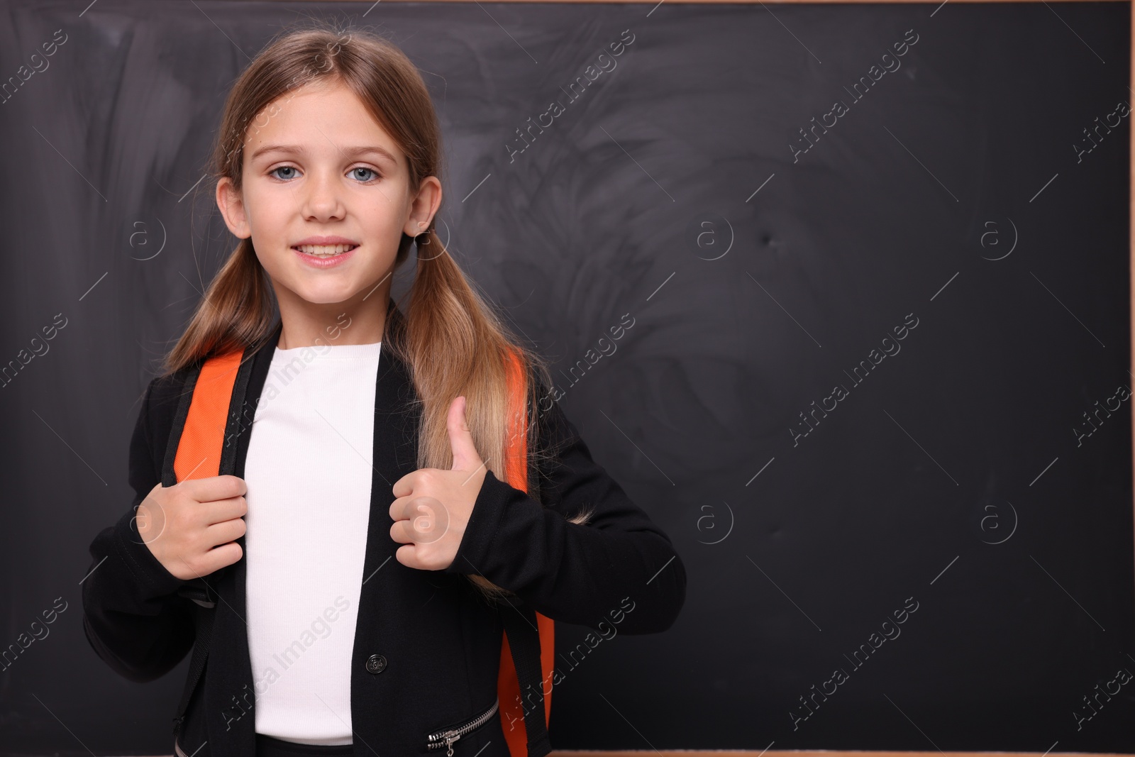 Photo of Smiling schoolgirl showing thumb up near blackboard. Space for text