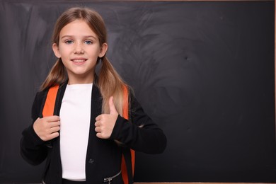 Photo of Smiling schoolgirl showing thumb up near blackboard. Space for text