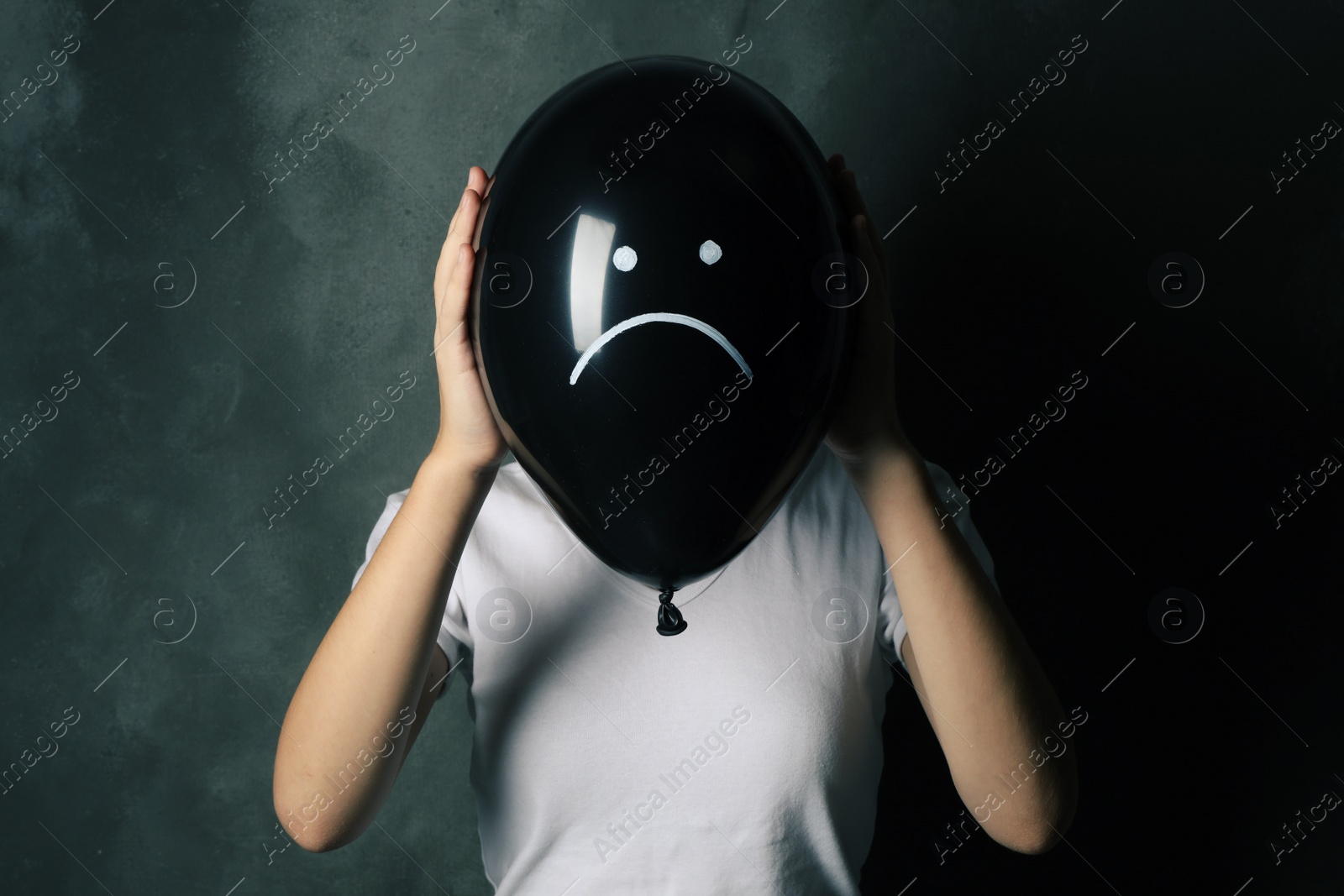 Photo of Woman hiding behind black balloon with drawn sad face on dark background. Depression symptoms