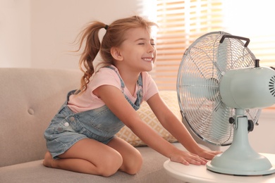 Photo of Little girl enjoying air flow from fan on sofa in living room. Summer heat