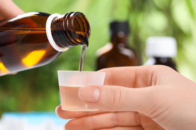 Photo of Woman pouring syrup from bottle into measuring cup on blurred background, closeup. Cold medicine