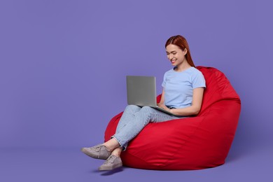 Photo of Smiling young woman working with laptop on beanbag chair against lilac background, space for text