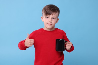 Photo of Cute boy with black ceramic mug showing thumbs up on light blue background