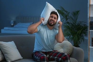 Photo of Unhappy young man covering ears with pillow in living room at night. Noisy neighbours