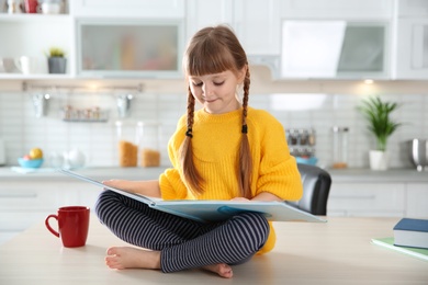Photo of Cute little girl reading book in kitchen at home