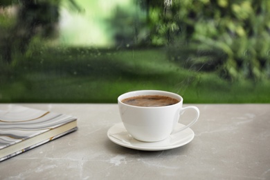 Photo of Cup of hot drink and notebook on stone windowsill against glass with rain drops, space for text