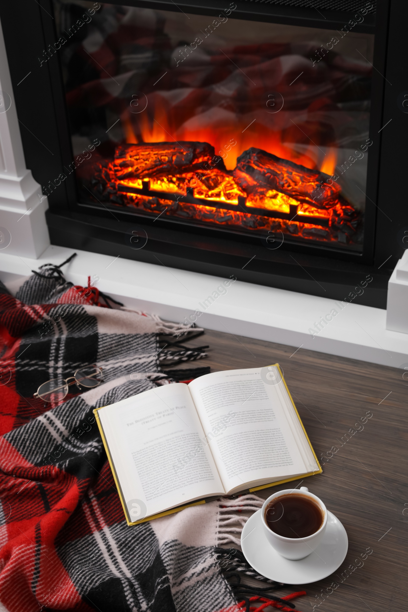 Photo of Cup of hot coffee, plaid and book on floor near fireplace, above view. Cozy atmosphere