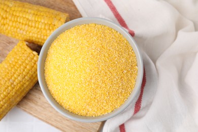 Photo of Raw cornmeal in bowl and corn cobs on white table, top view