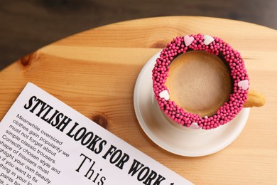 Photo of Delicious edible biscuit cupcoffee decorated with sprinkles and newspaper on wooden table, flat lay