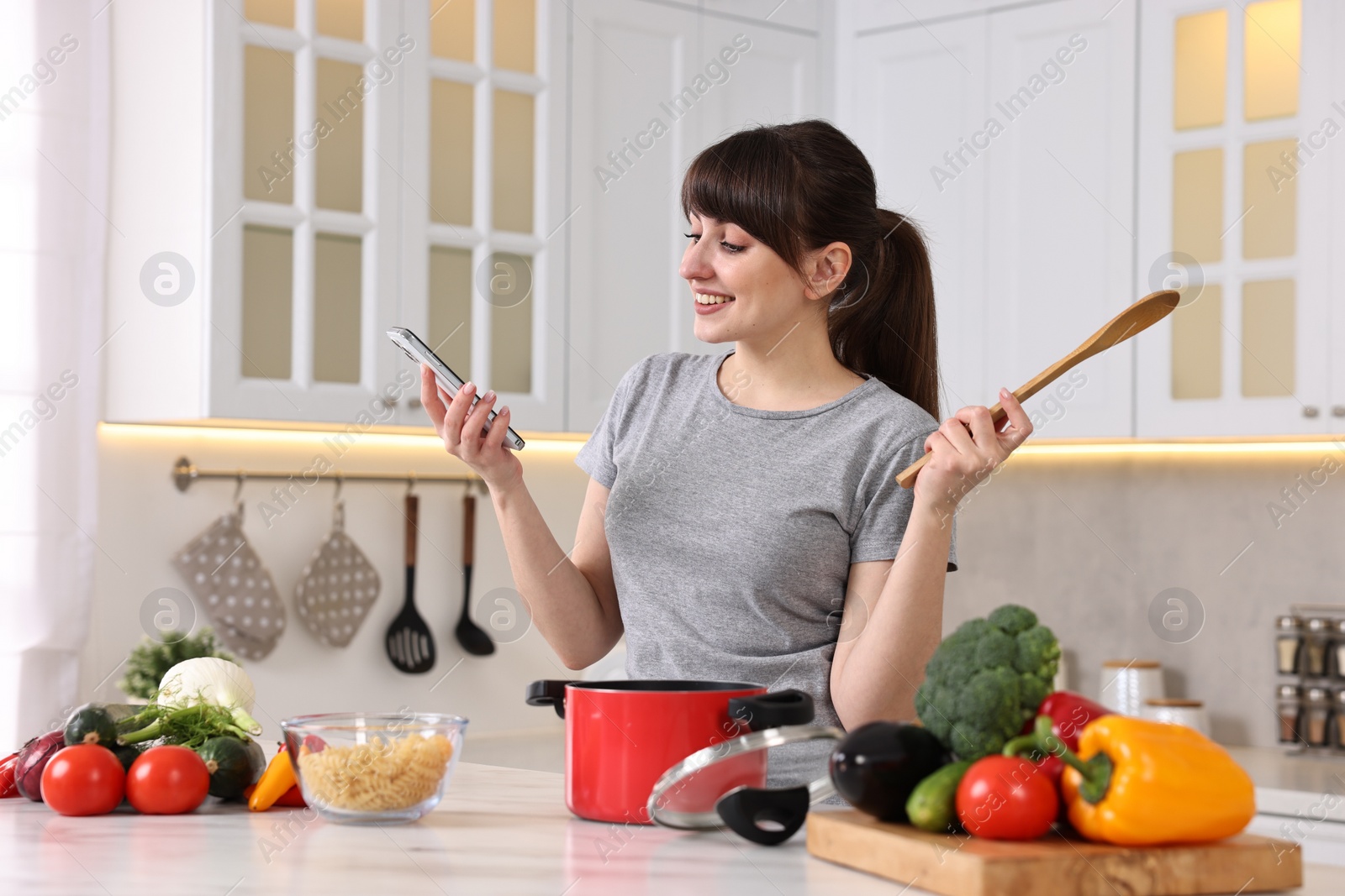Photo of Happy young housewife with spoon using smartphone while cooking at white marble table in kitchen