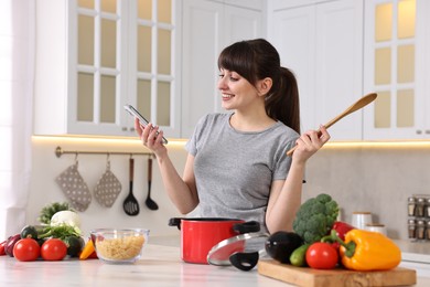 Photo of Happy young housewife with spoon using smartphone while cooking at white marble table in kitchen