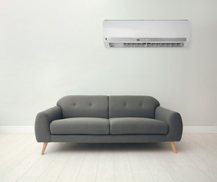 Modern air conditioner on white wall in room with stylish grey sofa