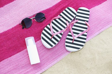 Towel with stylish flip flops, sunscreen and sunglasses on sandy beach, flat lay