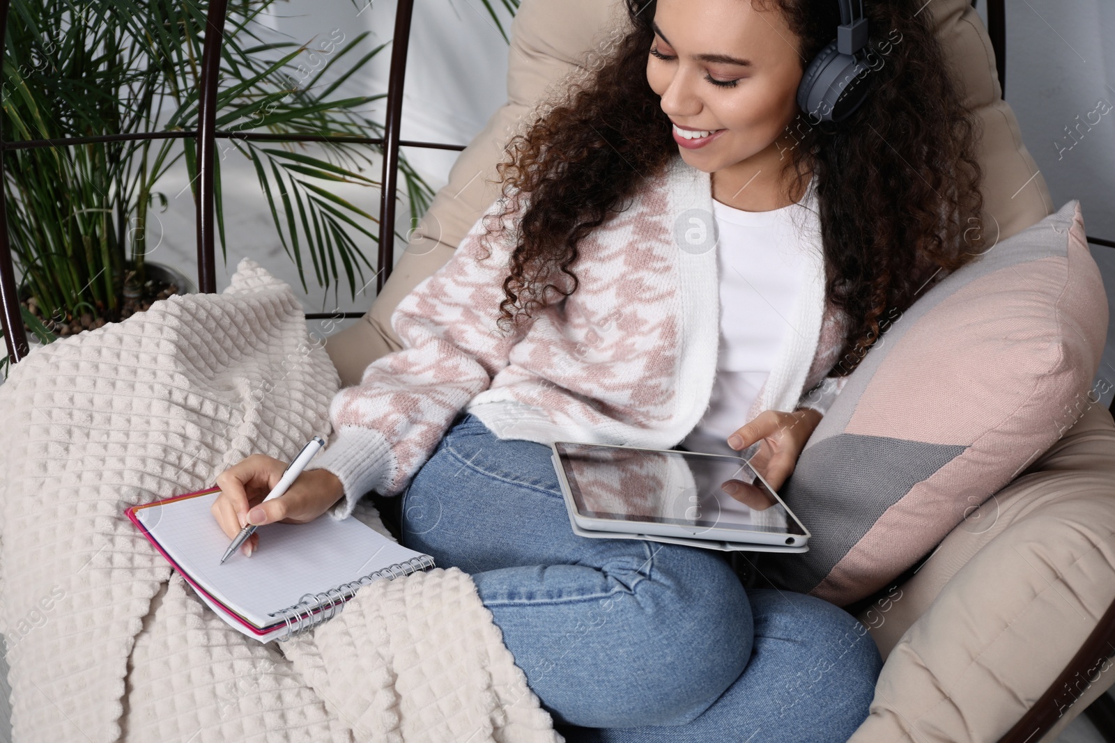 Photo of African American woman with headphones and tablet studying in egg chair at home. Distance learning