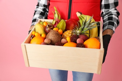 Courier holding crate with assortment of exotic fruits on pink background, closeup