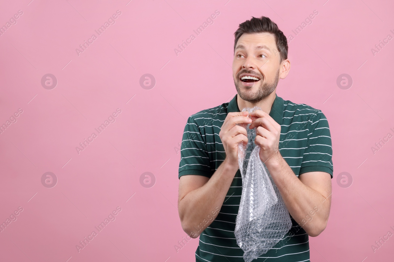 Photo of Happy man popping bubble wrap on pink background, space for text. Stress relief