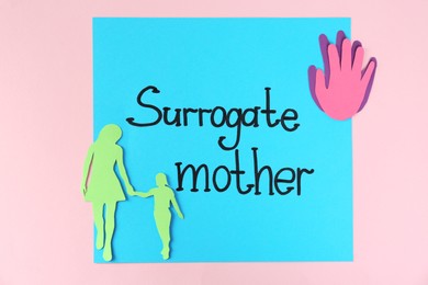 Photo of Note with words Surrogate Mother and paper cutouts on pink background, top view