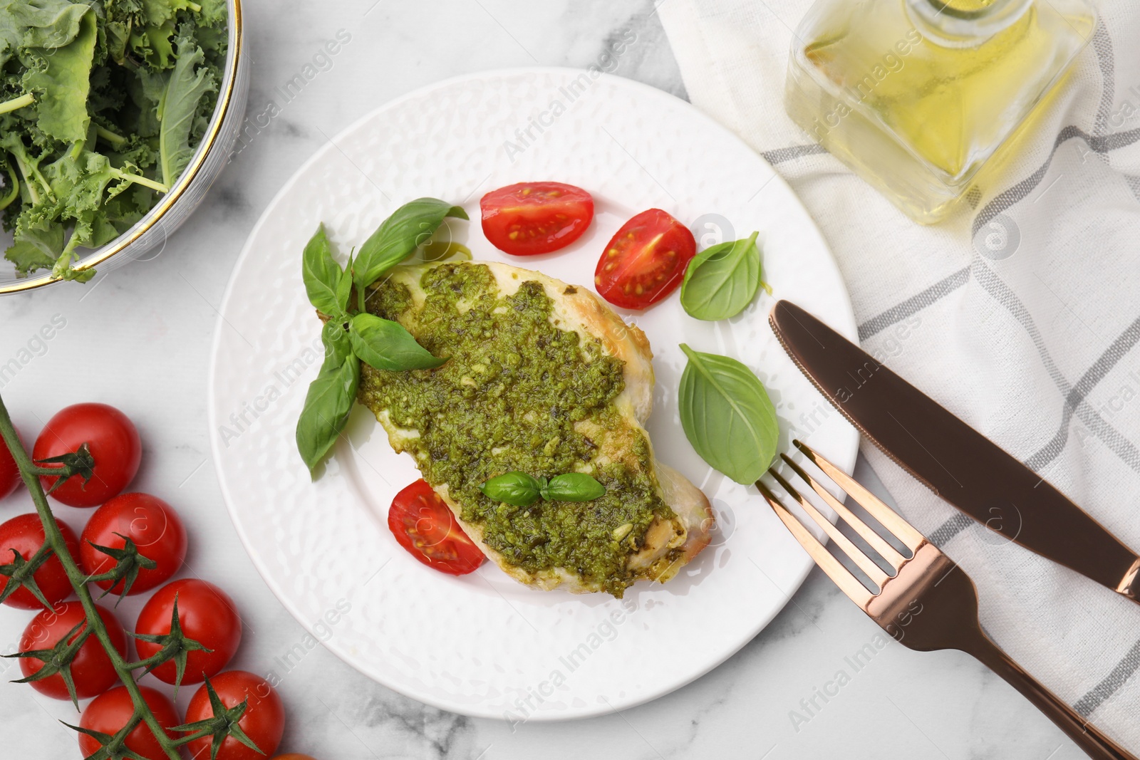 Photo of Delicious chicken breast with pesto sauce, tomatoes and cutlery served on white marble table, flat lay