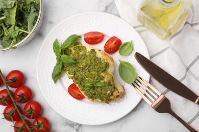 Photo of Delicious chicken breast with pesto sauce, tomatoes and cutlery served on white marble table, flat lay