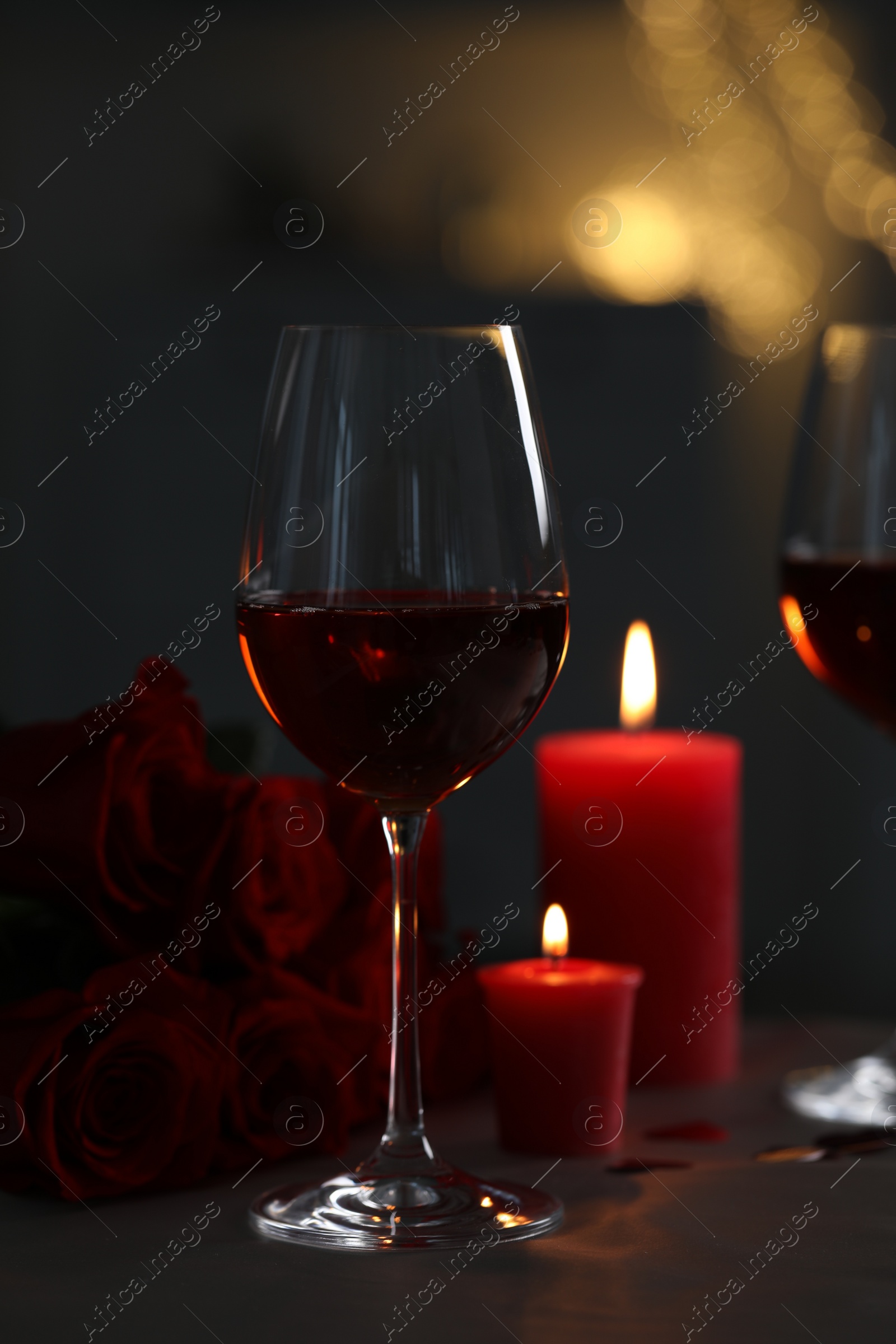 Photo of Glasses of red wine, burning candles and rose flowers on grey table against blurred lights. Romantic atmosphere