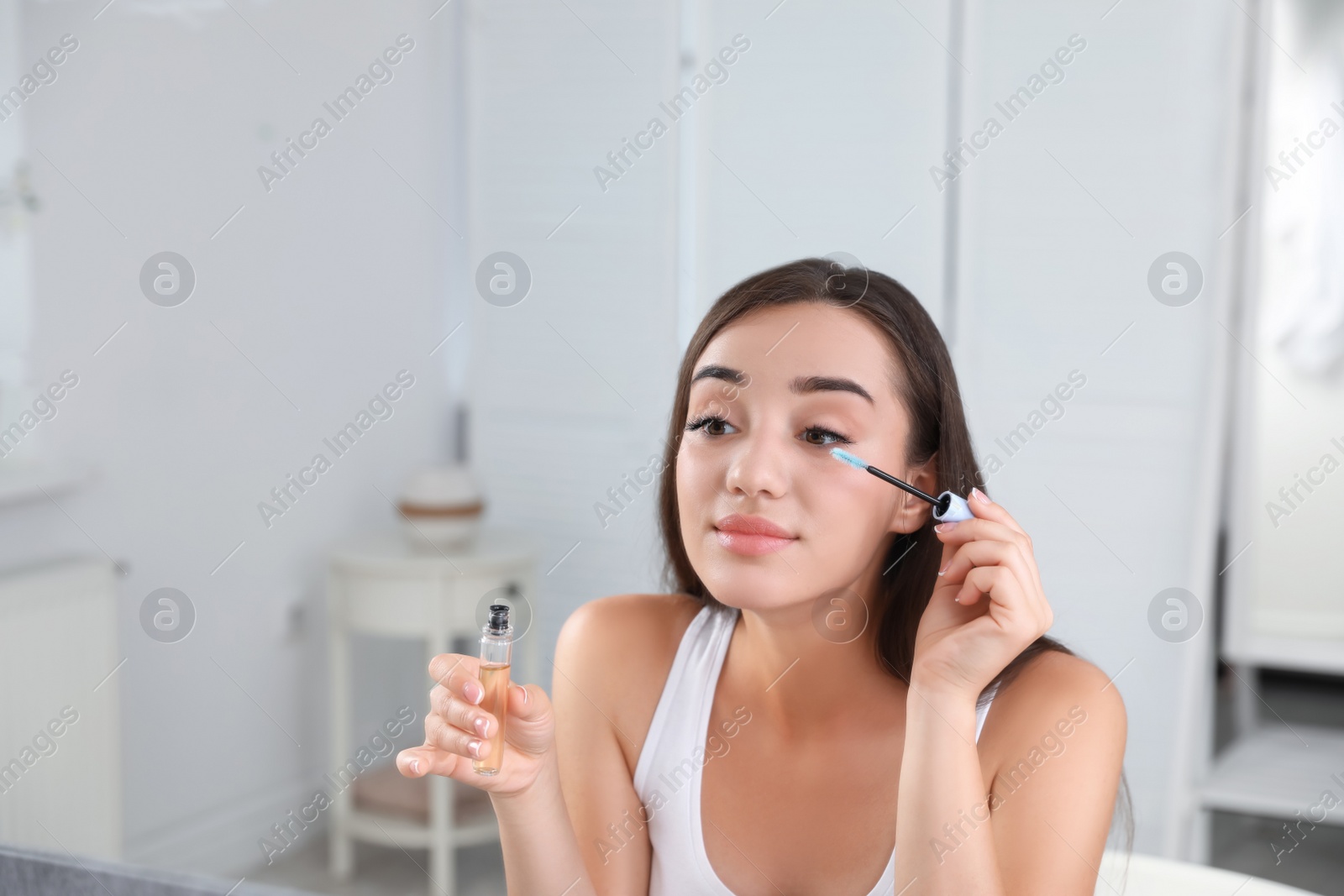 Photo of Young woman applying oil onto her eyelashes near mirror indoors