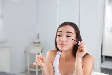 Young woman applying oil onto her eyelashes near mirror indoors