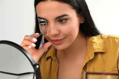 Photo of Beautiful young woman using eyelash curler in front of mirror indoors, closeup