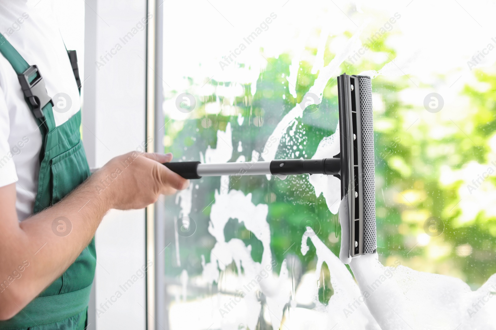 Photo of Male cleaner wiping window glass with squeegee indoors, closeup