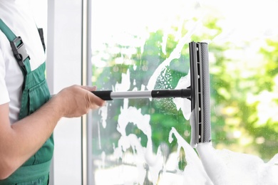 Male cleaner wiping window glass with squeegee indoors, closeup