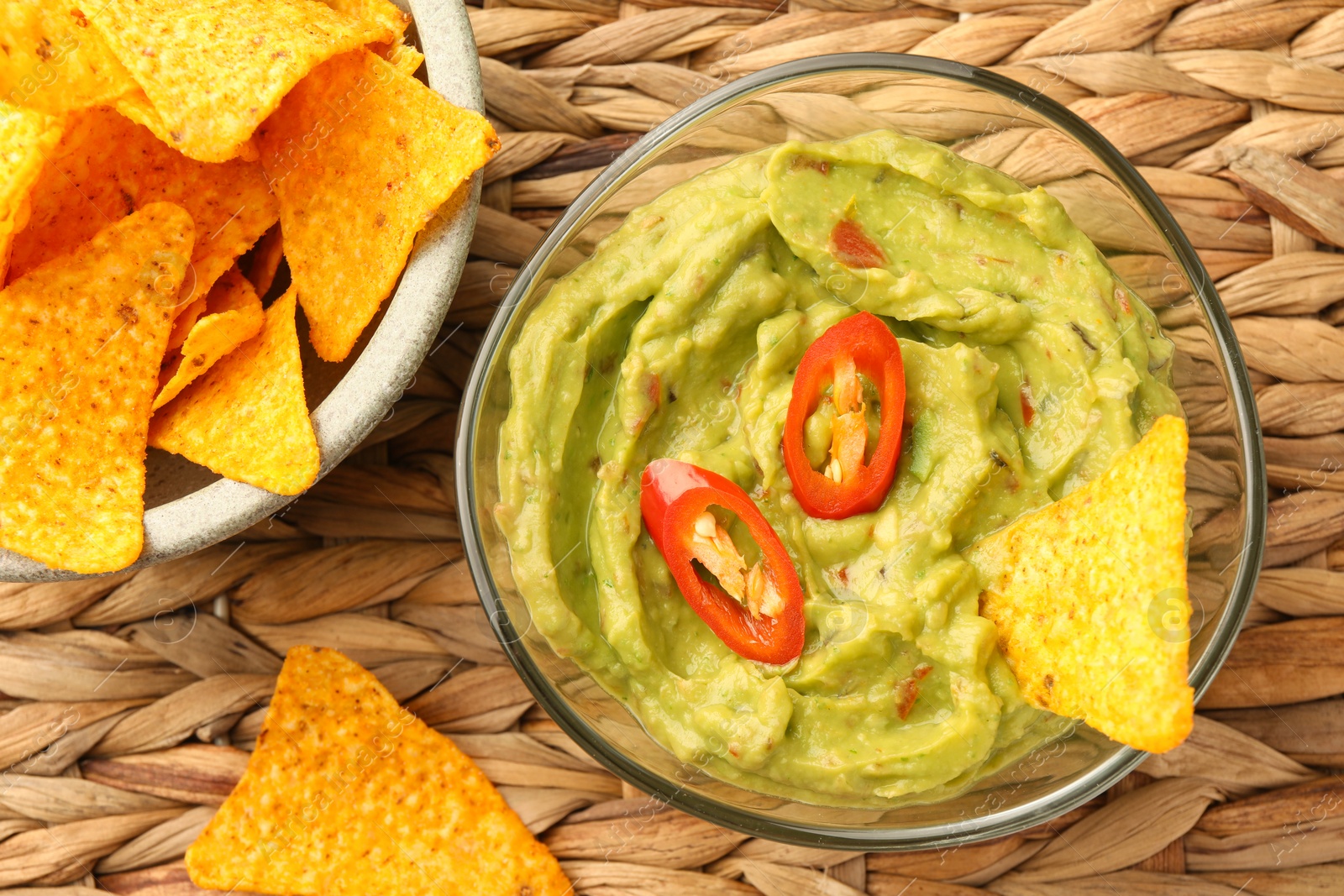 Photo of Bowl of delicious guacamole with chili pepper and nachos chips on wicker table, flat lay