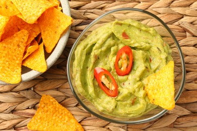 Bowl of delicious guacamole with chili pepper and nachos chips on wicker table, flat lay