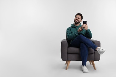 Happy young man using smartphone in armchair on white background, space for text