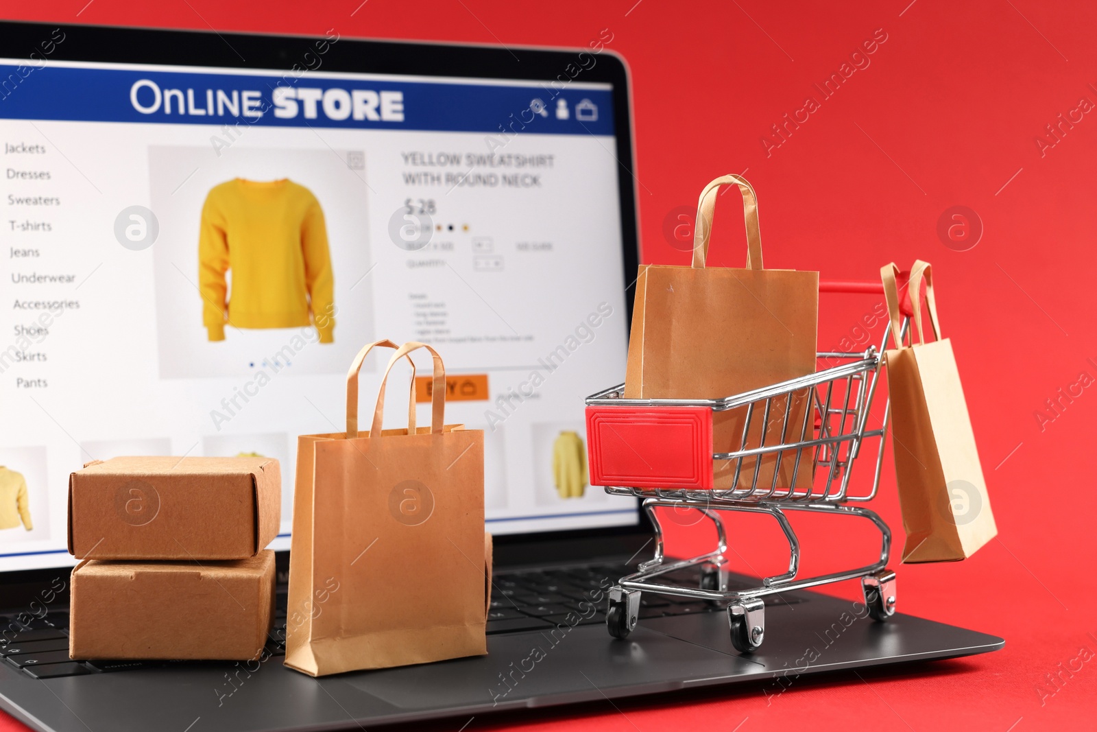 Photo of Online store. Laptop, mini shopping cart and small purchases on red background